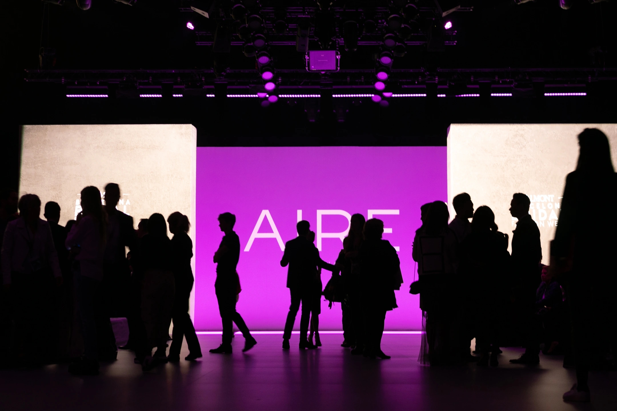 Minutos antes de empezar el desfile de AIRE Barcelona, cartel luminoso lila con el logotipo y personas enmedio del escenario en sombra. Pasarela de Valmont Barcelona Bridal Fashion Week.