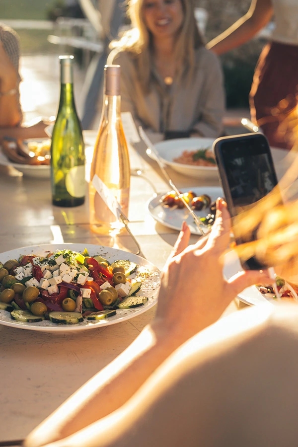 Fotografía de un plato de comida griego de queso feta con pimientos y una Mujer con copa de vino y botella de Viña Esmeralda mediterranean al atardecer con luz espectacular.